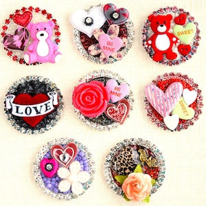 May include: A collection of decorative embellishments with rhinestone borders. Designs include a pink teddy bear, hearts, roses, and the word "LOVE". Colors include red, pink, white, and black. These embellishments are suitable for crafts and jewelry making.