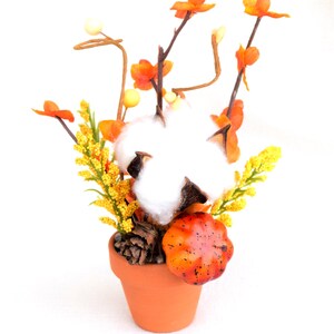 May include: A terracotta pot filled with a fall-themed arrangement. The arrangement includes a large cotton ball, orange and yellow flowers, a small pumpkin, and a pine cone. The background is white.
