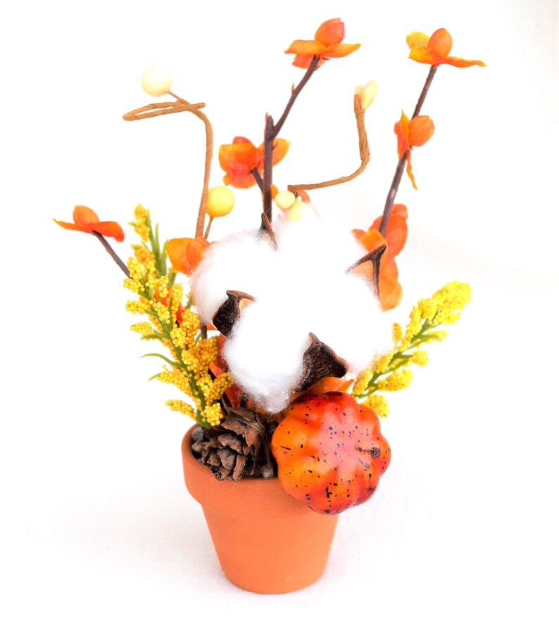May include: A terracotta pot filled with a fall-themed arrangement. The arrangement includes a large cotton ball, orange and yellow flowers, a small pumpkin, and a pine cone. The background is white.