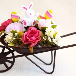 May include: A small metal wheelbarrow filled with pink and white flowers, a white bunny figurine, and pastel Easter eggs.