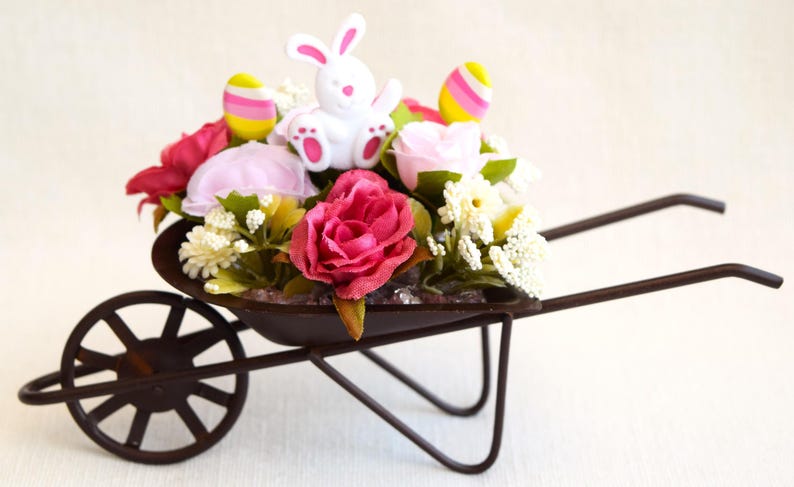 May include: A small metal wheelbarrow filled with pink and white flowers, a white bunny figurine, and pastel Easter eggs.