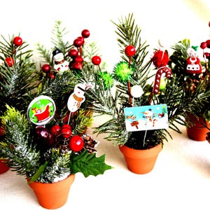 May include: Six small potted Christmas trees decorated with various ornaments, including a snowman, a gingerbread man, a candy cane, and a Santa Claus. The trees are in terracotta pots and are surrounded by a white background.
