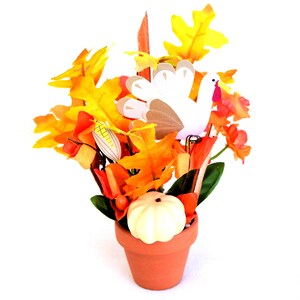 May include: Thanksgiving centerpiece in a terracotta pot. The arrangement features colorful fall leaves, a white pumpkin, a paper turkey, and decorative corn. The leaves are in shades of yellow, orange, and red. The pot is round and has a simple design.