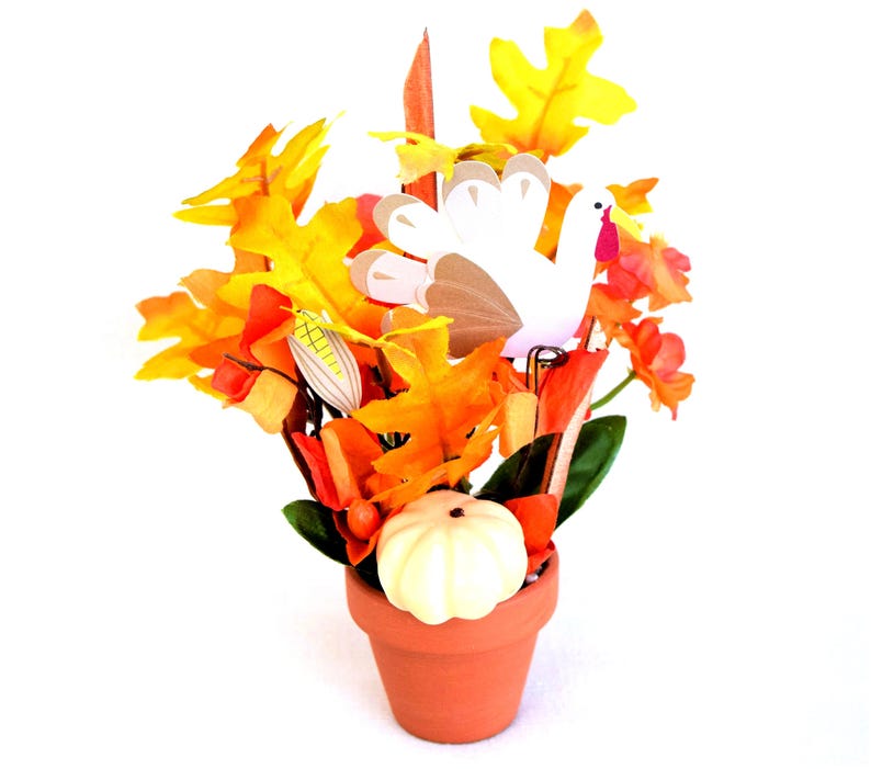 May include: Thanksgiving centerpiece in a terracotta pot. The arrangement features colorful fall leaves, a white pumpkin, a paper turkey, and decorative corn. The leaves are in shades of yellow, orange, and red. The pot is round and has a simple design.