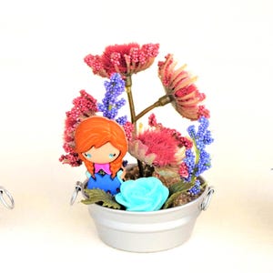 Pu&ograve; includere: Tre composizioni decorative in miniatura in piccoli secchi d'argento. Ognuna presenta una bambola con i capelli rossi e un vestito blu. Uno ha un albero in miniatura, un altro fiori e il terzo un mix di fiori e verde.
