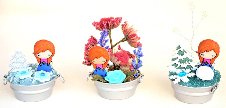 Pu&ograve; includere: Tre composizioni decorative in miniatura in piccoli secchi d'argento. Ognuna presenta una bambola con i capelli rossi e un vestito blu. Uno ha un albero in miniatura, un altro fiori e il terzo un mix di fiori e verde.