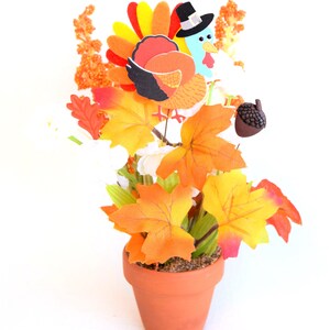 May include: Thanksgiving centerpiece in a terracotta pot. Features a cartoon turkey wearing a pilgrim hat, surrounded by orange and yellow fall leaves, white flowers, and an acorn. The arrangement is set against a white background.