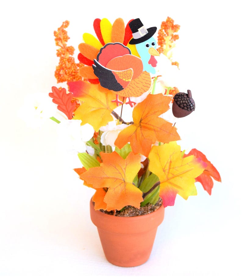 May include: Thanksgiving centerpiece in a terracotta pot. Features a cartoon turkey wearing a pilgrim hat, surrounded by orange and yellow fall leaves, white flowers, and an acorn. The arrangement is set against a white background.