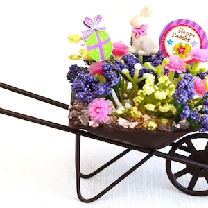 May include: A miniature wheelbarrow filled with purple, pink, and yellow flowers, a white bunny, and a green and purple Easter egg. The wheelbarrow is decorated with a sign that says "Happy Easter".