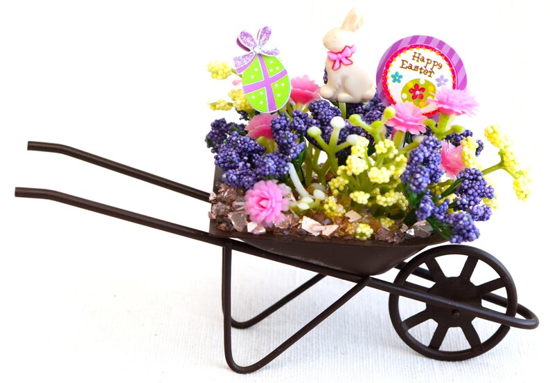 May include: A miniature wheelbarrow filled with purple, pink, and yellow flowers, a white bunny, and a green and purple Easter egg. The wheelbarrow is decorated with a sign that says "Happy Easter".