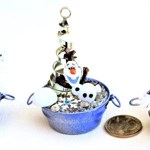 Pu&ograve; includere: Tre secchi blu in miniatura con manici, ciascuno contenente una piccola figura di Olaf, fiocchi di neve ed elementi decorativi. Un secchio ha una spirale argentata, un altro ha un piccolo albero argentato. Una moneta &egrave; accanto ai secchi.