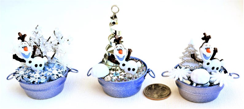 Pu&ograve; includere: Tre secchi blu in miniatura con manici, ciascuno contenente una piccola figura di Olaf, fiocchi di neve ed elementi decorativi. Un secchio ha una spirale argentata, un altro ha un piccolo albero argentato. Una moneta &egrave; accanto ai secchi.