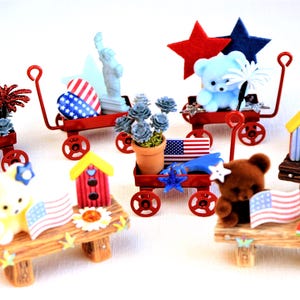 May include: A collection of miniature red wagons decorated with patriotic themes. Each wagon features a different American flag design, along with a variety of miniature figurines, including bears, a birdhouse, and a potted plant.