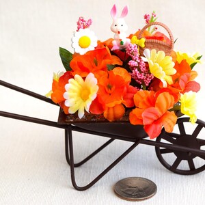 May include: A miniature brown metal wheelbarrow filled with orange, yellow, and white artificial flowers. A small white bunny with pink ears is sitting in the wheelbarrow.