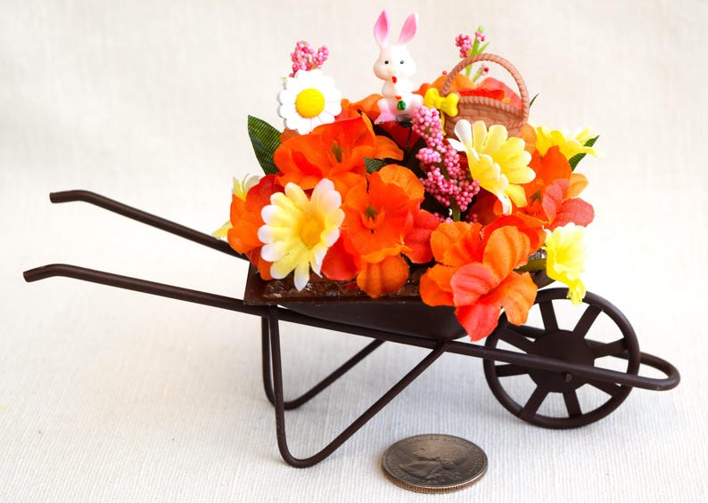 May include: A miniature brown metal wheelbarrow filled with orange, yellow, and white artificial flowers. A small white bunny with pink ears is sitting in the wheelbarrow.