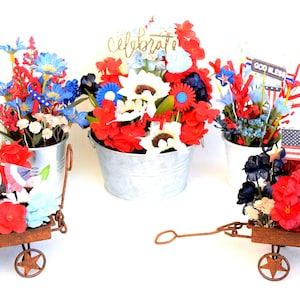 May include: Three miniature red, white, and blue flower arrangements in galvanized metal buckets are displayed on rustic metal wagons. The wagons have star-shaped wheels. The flower arrangements are decorated with patriotic themed embellishments, including a small American flag, a "God Bless" sign, and a "Celebrate" sign.