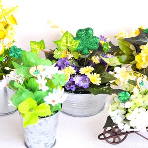 May include: A collection of small, galvanized metal containers filled with artificial flowers and foliage. The arrangements feature yellow, white, and purple flowers, shamrocks, and a leprechaun, celebrating St. Patrick's Day.