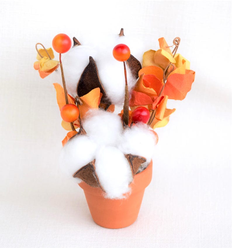 May include: A decorative arrangement of cotton bolls, orange berries, and fall-colored leaves in a small terracotta pot. The cotton bolls are fluffy and white, contrasting with the brown seed pods. The arrangement is set against a white background.