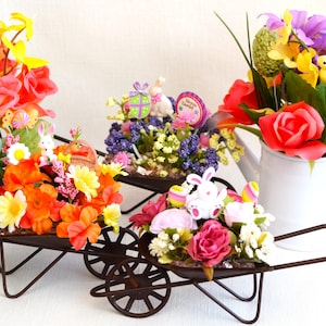 May include: Three miniature wheelbarrows filled with colorful artificial flowers and Easter decorations. The wheelbarrows are made of metal and have a rustic brown finish. The flowers include pink, orange, yellow, and purple roses, as well as white and purple flowers. The decorations include a bunny rabbit, a chick, and a small sign that says "Happy Easter".