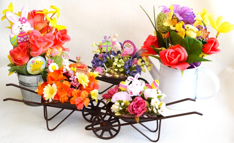 May include: Three miniature wheelbarrows filled with colorful artificial flowers and Easter decorations. The wheelbarrows are made of metal and have a rustic brown finish. The flowers include pink, orange, yellow, and purple roses, as well as white and purple flowers. The decorations include a bunny rabbit, a chick, and a small sign that says "Happy Easter".