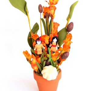 May include: A terracotta pot holds an arrangement of orange and green artificial flowers. Two small figurines dressed in Native American attire are nestled among the blooms. A white, scalloped decoration sits at the base of the arrangement.