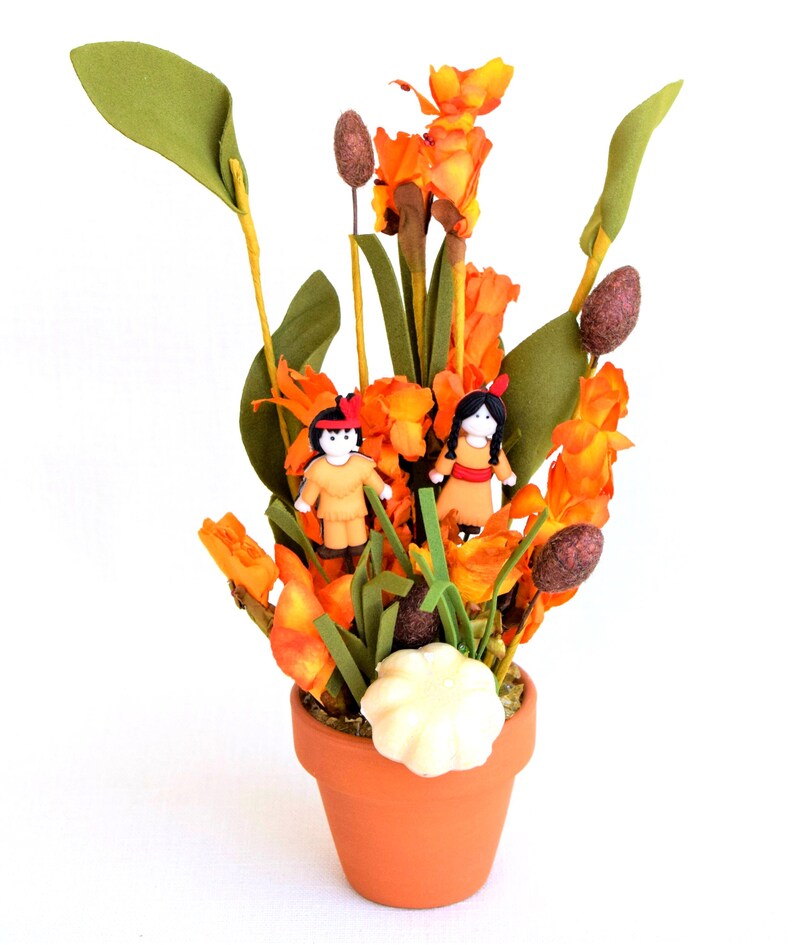 May include: A terracotta pot holds an arrangement of orange and green artificial flowers. Two small figurines dressed in Native American attire are nestled among the blooms. A white, scalloped decoration sits at the base of the arrangement.
