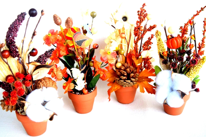 May include: Four small terracotta pots filled with fall-themed floral arrangements. Each arrangement features a variety of elements, including orange and yellow flowers, berries, cotton, and pinecones. The arrangements are set against a white background.