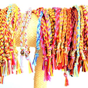 May include: Braided friendship bracelets in a variety of colors, including yellow, orange, red, pink, and blue. Each bracelet has a tassel and a decorative metal bead. The bracelets are displayed on a tan stand.