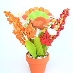 May include: A Thanksgiving-themed floral arrangement in a terracotta pot. The arrangement features a cartoon turkey, colorful leaves, and orange and red floral accents. The pot is filled with small white pebbles.