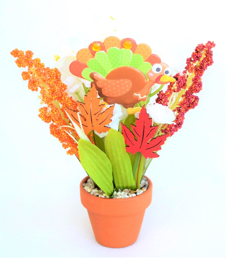 May include: A Thanksgiving-themed floral arrangement in a terracotta pot. The arrangement features a cartoon turkey, colorful leaves, and orange and red floral accents. The pot is filled with small white pebbles.
