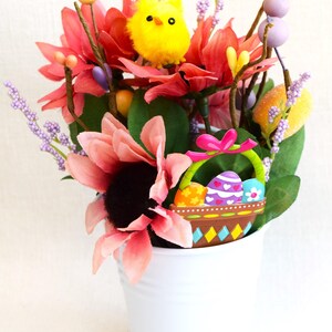 May include: A white metal bucket filled with a floral arrangement featuring pink, red, and yellow flowers, a yellow chick, and a basket with colorful Easter eggs.