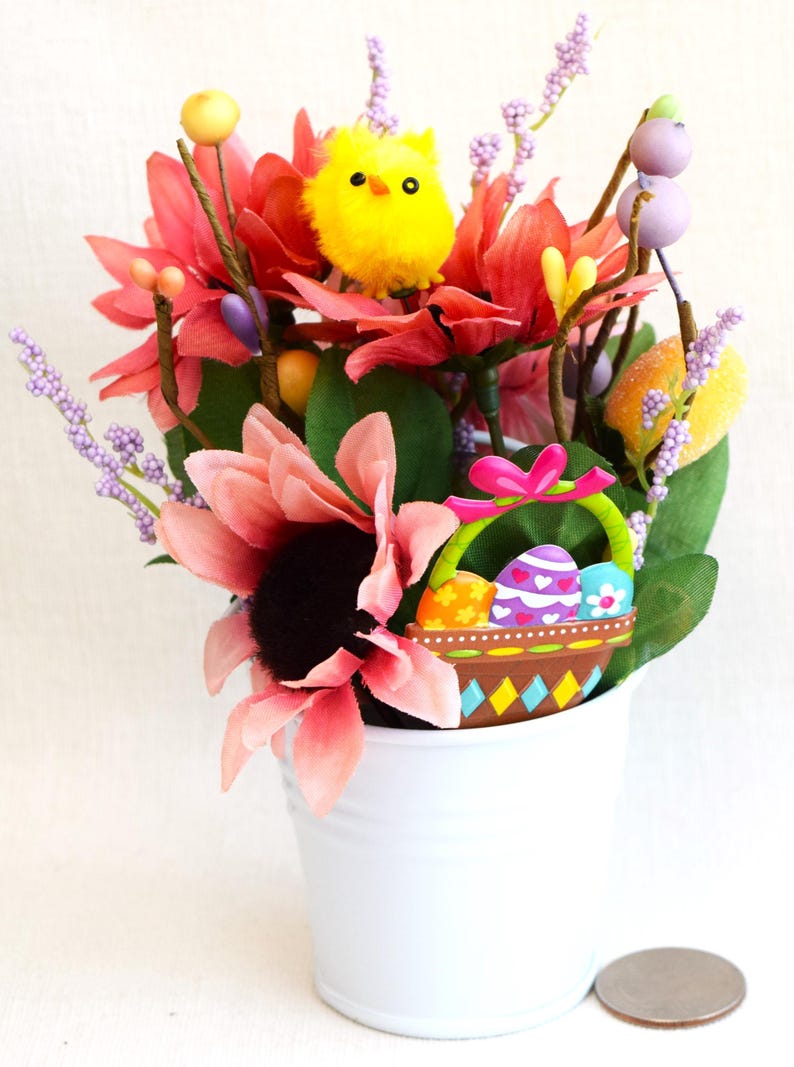 May include: A white metal bucket filled with a floral arrangement featuring pink, red, and yellow flowers, a yellow chick, and a basket with colorful Easter eggs.
