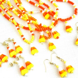May include: A collection of jewelry featuring candy corn charms, including several beaded necklaces with orange, yellow, and white seed beads, and multiple pairs of matching drop earrings with gold-toned hooks and candy corn-shaped pendants.