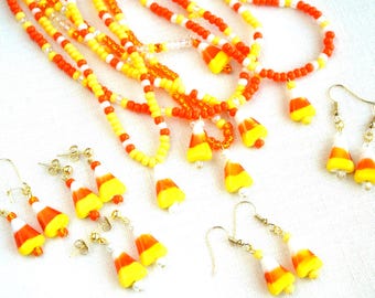 May include: A collection of jewelry featuring candy corn charms, including several beaded necklaces with orange, yellow, and white seed beads, and multiple pairs of matching drop earrings with gold-toned hooks and candy corn-shaped pendants.