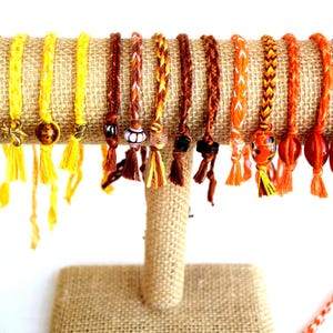 May include: A collection of fifteen braided cord bracelets in shades of yellow, orange, and brown, featuring various decorative beads, charms, and small tassels at the ends, displayed in a row on a cylindrical burlap-covered jewelry stand.