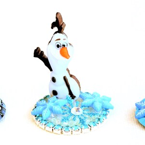 Pu&ograve; includere: Tre statuette di Olaf dal film Frozen. Ogni statuetta &egrave; bianca con braccia e capelli neri, un naso arancione e un'espressione sorridente. Sono poste su una base con dettagli di fiocchi di neve e ghiaccio blu.