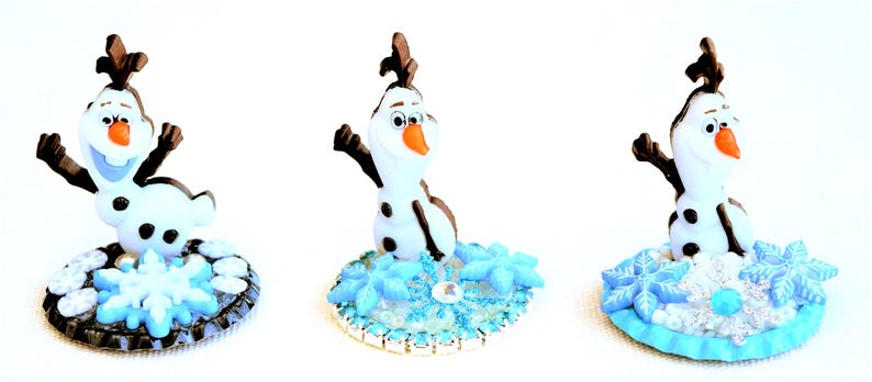 Pu&ograve; includere: Tre statuette di Olaf dal film Frozen. Ogni statuetta &egrave; bianca con braccia e capelli neri, un naso arancione e un'espressione sorridente. Sono poste su una base con dettagli di fiocchi di neve e ghiaccio blu.