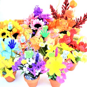 May include: A collection of small potted plants decorated with colorful school-themed items, including crayons, an eraser, and handprint cutouts. The plants are in terracotta pots and feature a variety of flowers, including sunflowers, daisies, and roses.