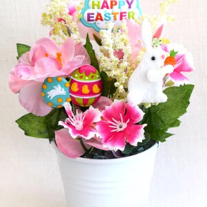 May include: A white metal bucket filled with pink and white flowers, green leaves, a white bunny holding a carrot, and colorful Easter eggs. The bucket has a "Happy Easter" sign.