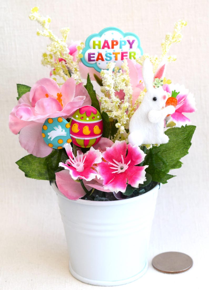 May include: A white metal bucket filled with pink and white flowers, green leaves, a white bunny holding a carrot, and colorful Easter eggs. The bucket has a "Happy Easter" sign.