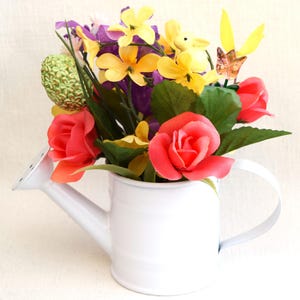 May include: A white metal watering can with a handle, filled with a colorful bouquet of artificial flowers. The flowers include yellow, pink, purple, and green.