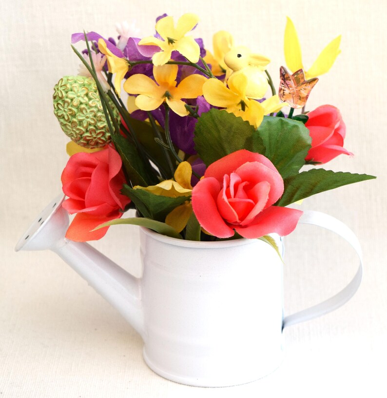 May include: A white metal watering can with a handle, filled with a colorful bouquet of artificial flowers. The flowers include yellow, pink, purple, and green.
