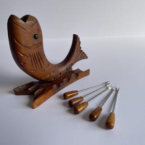 May include: A wooden fish-shaped serving set with a carved design. The set includes a fish-shaped holder and five small forks with wooden handles and metal tines. The wood has a warm, brown tone.