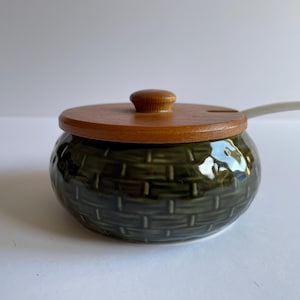 May include: A green ceramic sugar bowl with a woven pattern and a wooden lid. The bowl has a small white spoon attached to the lid.