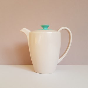 May include: A white ceramic teapot with a teal lid and handle. The teapot has a rounded body and a spout on the side.