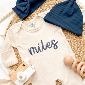 Peut inclure: Ensemble de vêtements pour bébé comprenant un body crème brodé du nom "Miles" en bleu marine. Comprend un bandeau et un bonnet bleu marine, un appareil photo jouet en bois et un hochet en bois. Le texte "Explore Daisy" est également visible.