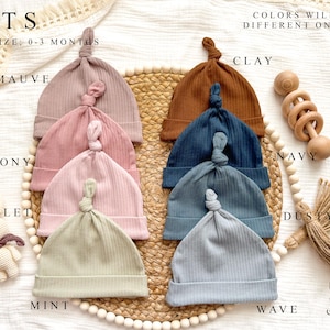Peut inclure: Collection de bonnets pour b&eacute;b&eacute;s en tricot c&ocirc;tel&eacute;, disponibles en plusieurs couleurs, dont mauve, pivoine et menthe. Les bonnets sont pr&eacute;sent&eacute;s sur un tapis tress&eacute;, avec un hochet en bois et un petit jouet. Le texte indique "HATS" et "ONLY SIZE: 0-3 MONTHS".