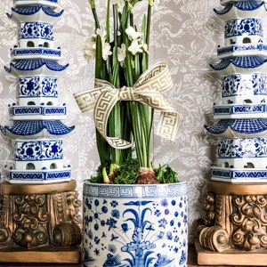 May include: White paperwhites in bloom with green leaves are arranged in a blue and white ceramic planter with a gold ribbon. The planter is decorated with a floral design and a geometric border. The flowers are displayed on a wooden surface with two blue and white ceramic pagodas on either side.