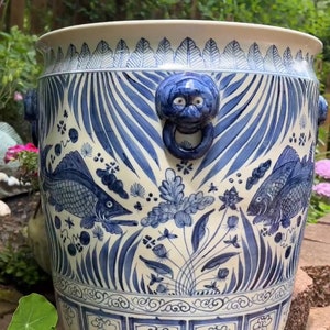 May include: A large, white ceramic planter with blue and white floral and fish designs. The planter has two blue and white lion head handles with rings.
