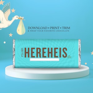 May include: A light blue candy bar wrapper with the text "HEREHEIS" in brown letters, with the words "IT'S A BOY!" on the side. The wrapper is on a white pedestal, with a stork illustration in the background.